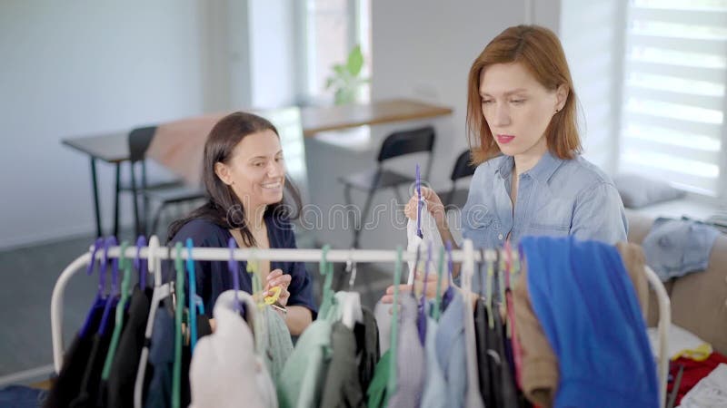 Één vrouw zet kleding op hanger in ruimte en haar vrouwelijke vriend is scherpe draden met grappig gezicht