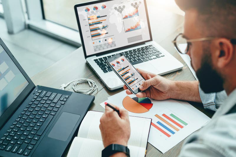 întreprinzătorul stă în birou utilizează smartphone-ul care lucrează pe laptop cu diagrame pe ecran care face note în notebook royalty free stock photography