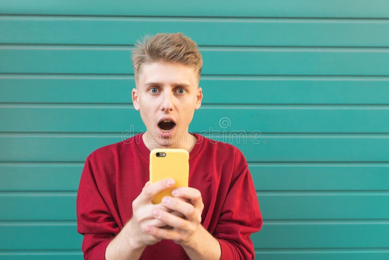 Έκπληκτος νεαρός άνδρας με ένα smartphone στα χέρια του που εξετάζουν τη κάμερα ενάντια στο σκηνικό ενός τυρκουάζ τοίχου στοκ εικόνες Έκπληκτος νεαρός άνδρας με ένα smartphone στα χέρια του που εξετάζουν τη κάμερα ενάντια στο σκηνικό ενός τυρκουάζ τοίχου στοκ εικόνες