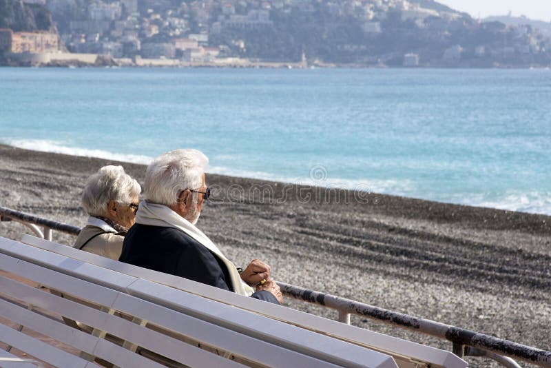 Ένα ώριμο, γκρίζος-μαλλιαρό όμορφο ζεύγος: ένας άνδρας και μια γυναίκα κάθονται σε έναν άσπρο πάγκο στον περίπατο des Anglais και στοκ φωτογραφίες Ένα ώριμο, γκρίζος-μαλλιαρό όμορφο ζεύγος: ένας άνδρας και μια γυναίκα κάθονται σε έναν άσπρο πάγκο στον περίπατο des Anglais και στοκ φωτογραφίες