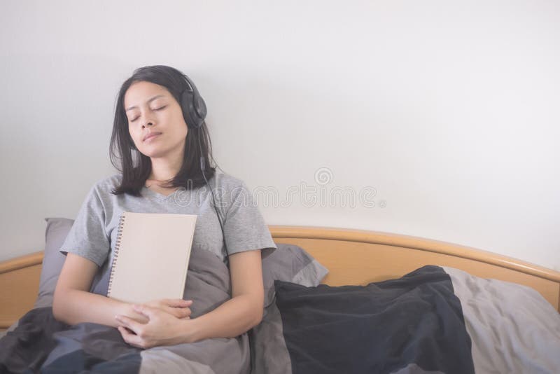 Όμορφη ασιατική μουσική ακούσματος γυναικών με το ακουστικό και ανάγνωση της χαλάρωσης βιβλίων στο κρεβάτι στοκ φωτογραφία με δικαίωμα ελεύθερης χρήσης Όμορφη ασιατική μουσική ακούσματος γυναικών με το ακουστικό και ανάγνωση της χαλάρωσης βιβλίων στο κρεβάτι στοκ φωτογραφία με δικαίωμα ελεύθερης χρήσης