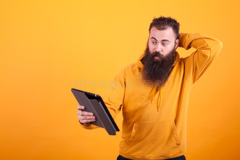 Όμορφος νεαρός άνδρας με τη μακριά γενειάδα που εξετάζει συγκεχυμένος την ταμπλέτα πέρα από το κίτρινο υπόβαθρο στοκ εικόνες με δικαίωμα ελεύθερης χρήσης Όμορφος νεαρός άνδρας με τη μακριά γενειάδα που εξετάζει συγκεχυμένος την ταμπλέτα πέρα από το κίτρινο υπόβαθρο στοκ εικόνες με δικαίωμα ελεύθερης χρήσης
