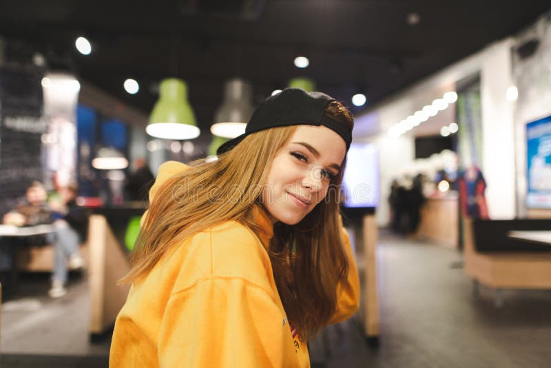 Το πρότυπο σε μια ΚΑΠ και έναν πορτοκαλή hoodie θέτει στη κάμερα στο εσωτερικό, εξετάζει τη κάμερα και χαμογελά στοκ φωτογραφία Το πρότυπο σε μια ΚΑΠ και έναν πορτοκαλή hoodie θέτει στη κάμερα στο εσωτερικό, εξετάζει τη κάμερα και χαμογελά στοκ φωτογραφία
