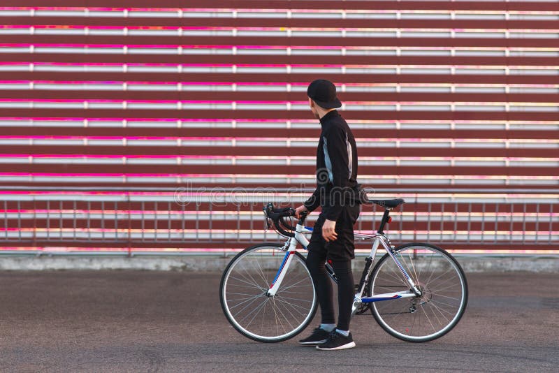 Ποδηλάτης σκοτεινό sportswear, που στέκεται με ένα ποδήλατο στο υπόβαθρο μιας κόκκινης οθόνης και που εξετάζει το στοκ φωτογραφία με δικαίωμα ελεύθερης χρήσης Ποδηλάτης σκοτεινό sportswear, που στέκεται με ένα ποδήλατο στο υπόβαθρο μιας κόκκινης οθόνης και που εξετάζει το στοκ φωτογραφία με δικαίωμα ελεύθερης χρήσης