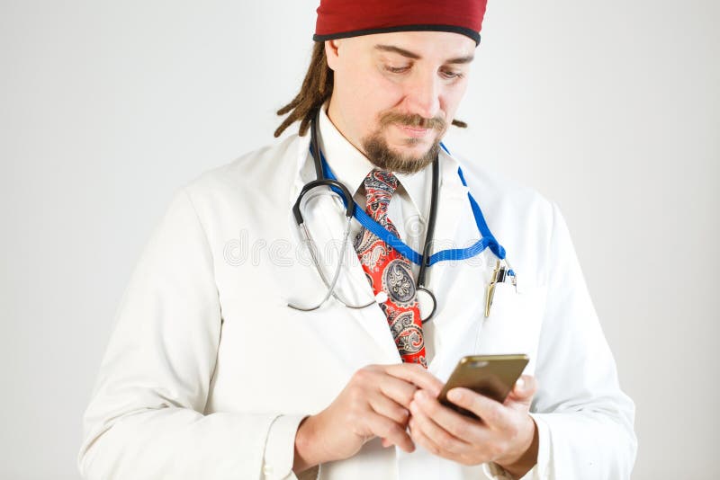Доктор с dreadlocks и бородой держит смартфон в его руках, стетоскоп и значок висит вокруг его шею стоковая фотография rf Доктор с dreadlocks и бородой держит смартфон в его руках, стетоскоп и значок висит вокруг его шею стоковая фотография rf