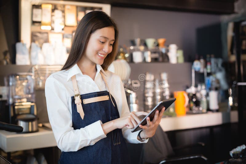 Азиатское barista женщины усмехаясь с таблеткой в ее руке, женские работники принимает заказы от онлайн клиентов стоковая фотография rf Азиатское barista женщины усмехаясь с таблеткой в ее руке, женские работники принимает заказы от онлайн клиентов стоковая фотография rf