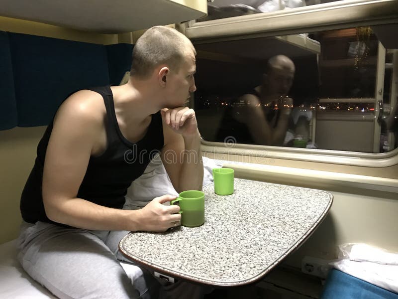 Кавказский молодой человек сидит на таблице в вагоне, держит зеленую чашку чаю и смотрит внимательно вне окно на светах города но стоковые фото Кавказский молодой человек сидит на таблице в вагоне, держит зеленую чашку чаю и смотрит внимательно вне окно на светах города но стоковые фото
