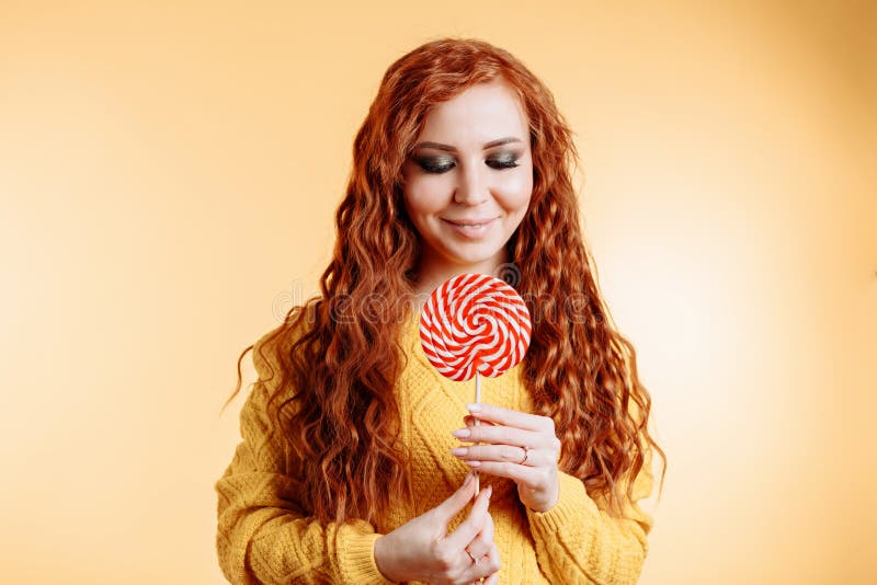 Красивая молодая курчавая женщина redhead держа красный белый леденец на палочке и смотря вниз стоковая фотография Красивая молодая курчавая женщина redhead держа красный белый леденец на палочке и смотря вниз стоковая фотография