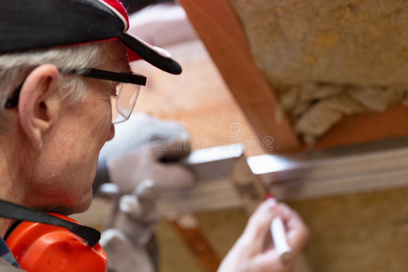 Man in googles making a mark on metal frame on unfinished attic ceiling. Attic insulation and renovation in natural light. Man in googles making a mark on metal frame on unfinished attic ceiling. Attic insulation and renovation in natural light