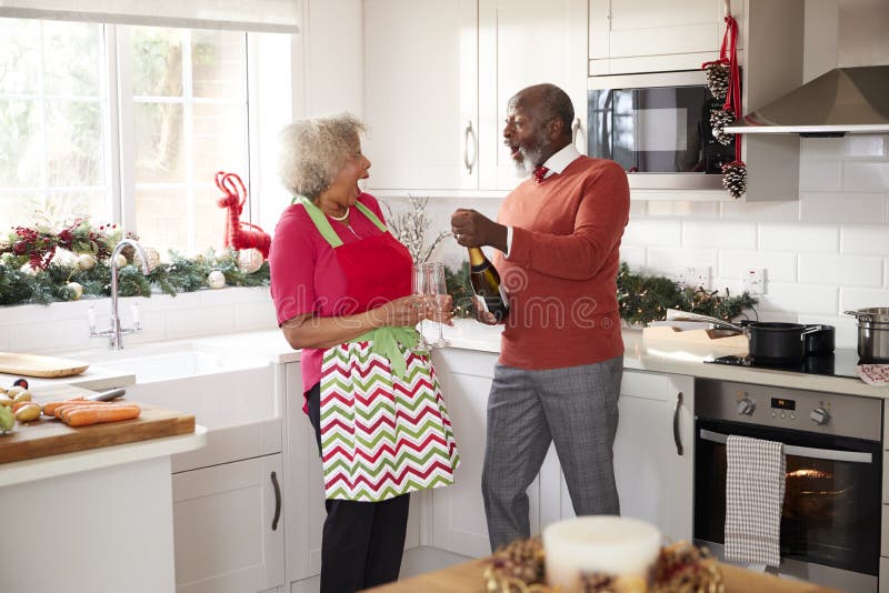 Öffnender Champagner der älteren schwarzen Paare und zusammen lachen in der Küche beim Vorbereiten des Abendessens am Weihnachtst lizenzfreie stockfotografie Öffnender Champagner der älteren schwarzen Paare und zusammen lachen in der Küche beim Vorbereiten des Abendessens am Weihnachtst lizenzfreie stockfotografie