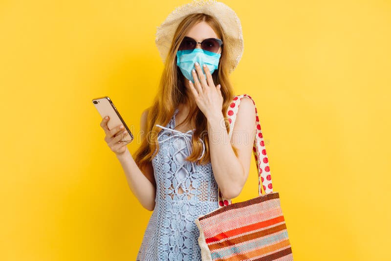 överraskad ung kvinna i medicinsk mask i sommarklänning och som använder mobiltelefon över gul bakgrund royaltyfria foton