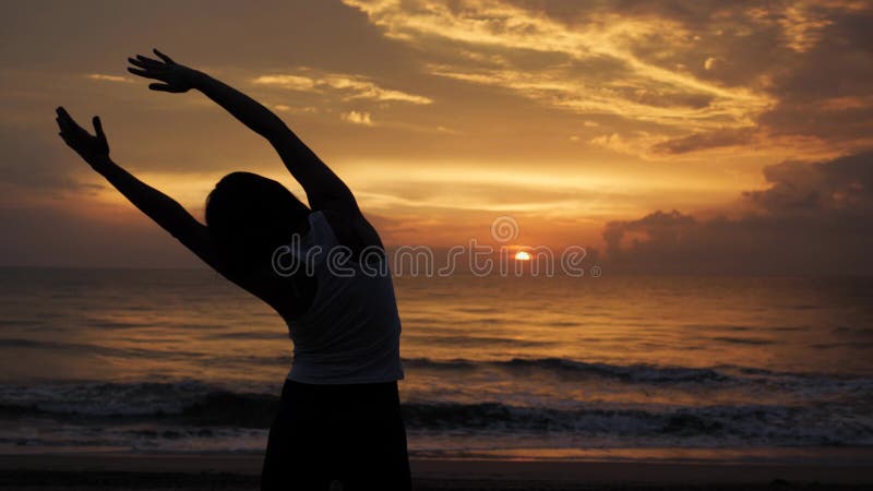 Übendes Yoga der jungen gesunden Frau auf dem Strand an den Sonnenuntergangmeereswogen Starke Vertrauensfrau unter dem Sonnenunte