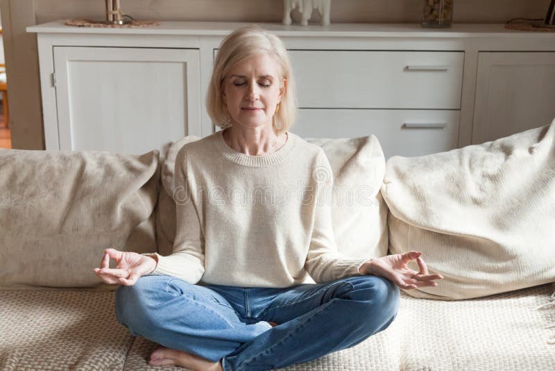 Calm senior woman relax on couch meditating in lotus position, peaceful aged female practice yoga at home relieve stress or renew energy, elderly lady do physical exercises following healthy lifestyle. Calm senior woman relax on couch meditating in lotus position, peaceful aged female practice yoga at home relieve stress or renew energy, elderly lady do physical exercises following healthy lifestyle