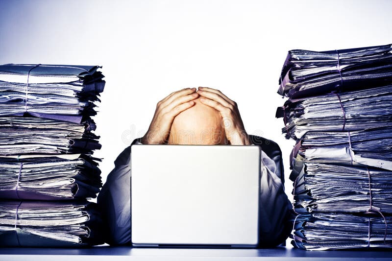 Male businessman sitting behind a laptop, his face hidden, with his hands on top of his head. Two large piles of paperwork are piled on each side of the model, towering over his head. Isolated on white background. Male businessman sitting behind a laptop, his face hidden, with his hands on top of his head. Two large piles of paperwork are piled on each side of the model, towering over his head. Isolated on white background.