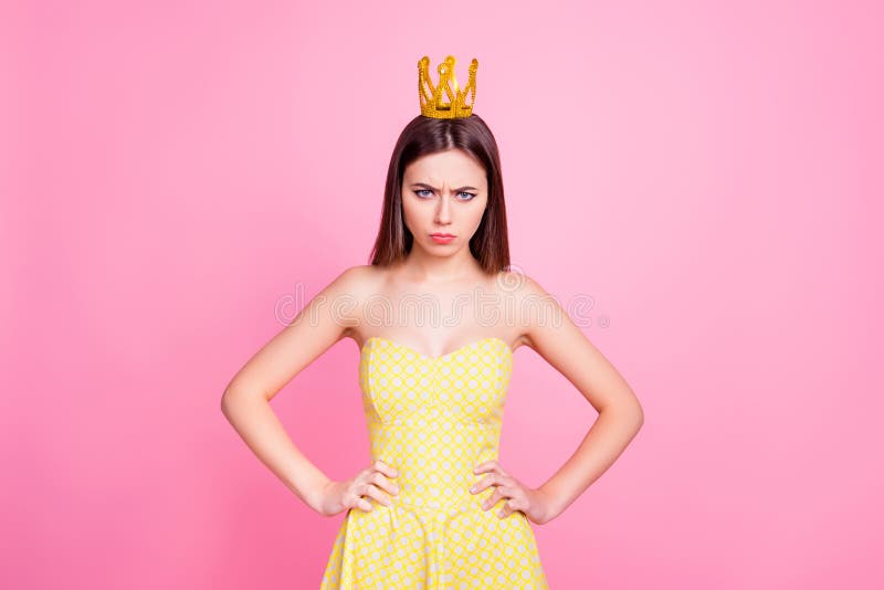 Confident offended persistent formidable woman looking at camera with angry gaze holding arms at hips, having gold crown on her head, wearing yellow dress, isolated on pink background. Confident offended persistent formidable woman looking at camera with angry gaze holding arms at hips, having gold crown on her head, wearing yellow dress, isolated on pink background