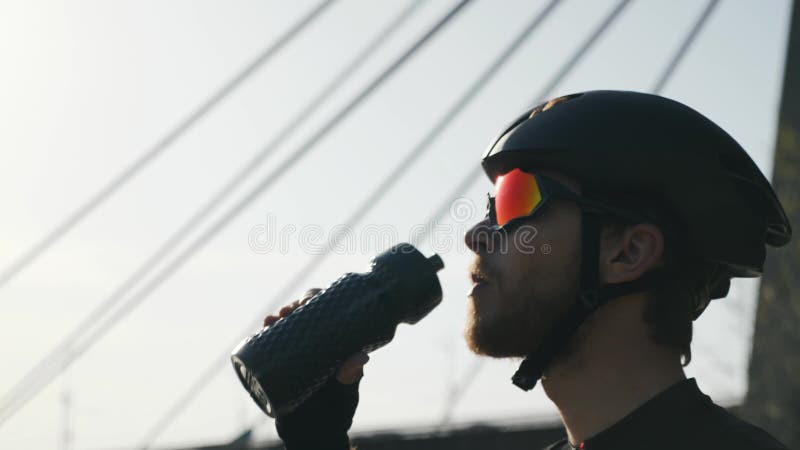 Überzeugter Radfahrer mit Trinkwasser des roten Bartes von tragendem Sturzhelm und Sonnenbrille der Flasche Brücke und Fluss im H