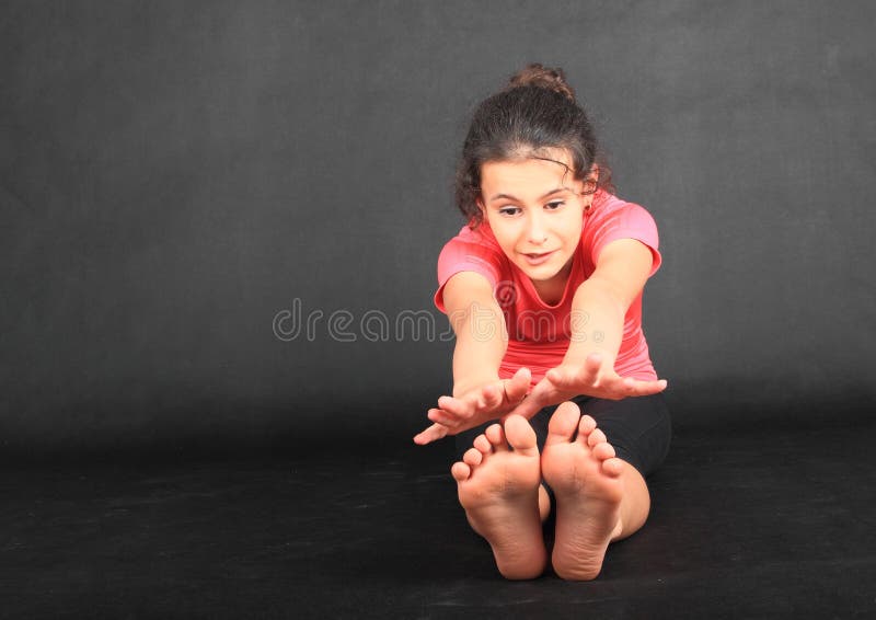 Pretty girl - barefoot brunette schoolkid dressed in pink t-shirt, exercising yoga pose seated forward fold or paschimottanasana with hands trying to reach soles of bare feet. Active childhood concept. Pretty girl - barefoot brunette schoolkid dressed in pink t-shirt, exercising yoga pose seated forward fold or paschimottanasana with hands trying to reach soles of bare feet. Active childhood concept.