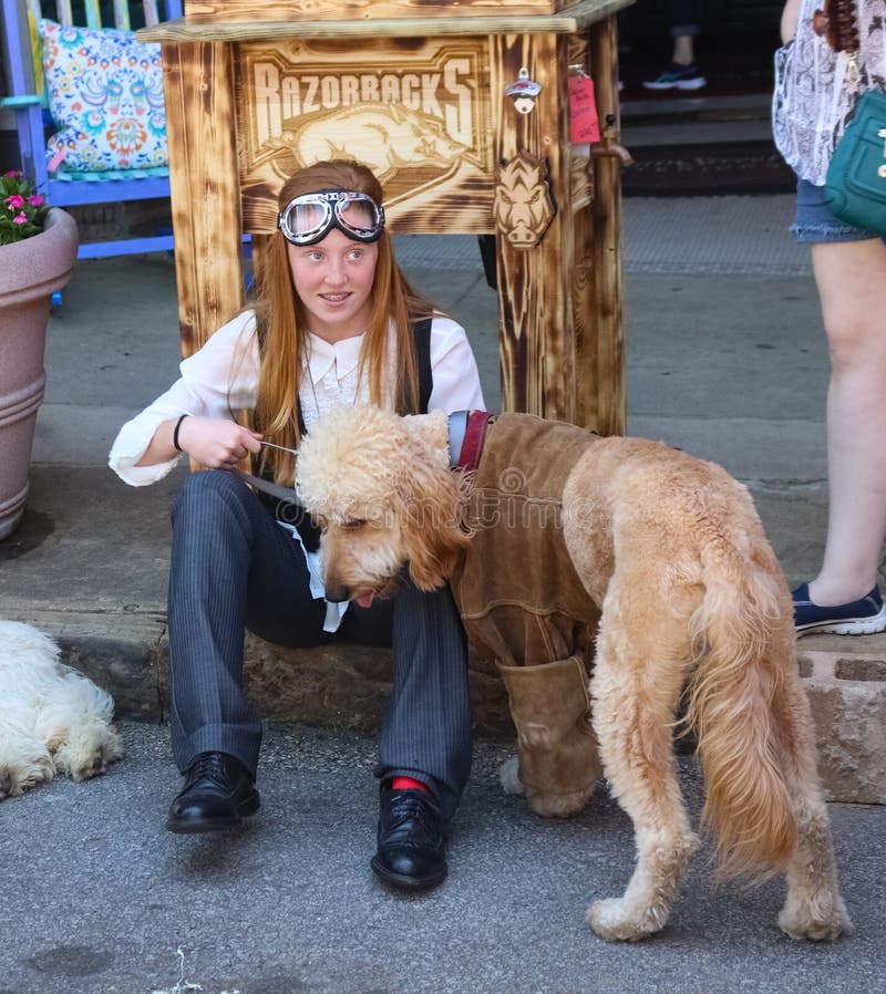 Ładna młoda dziewczyna z brasami i długim czerwonym włosy z steampunk gogle siedzi na krawężniku z labradoodle psem ubierającym w obraz royalty free Ładna młoda dziewczyna z brasami i długim czerwonym włosy z steampunk gogle siedzi na krawężniku z labradoodle psem ubierającym w obraz royalty free