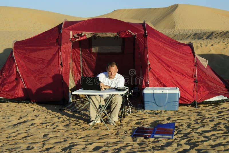 Man with a laptop and portable folding solar kit charger in front of his tent in the sandy desert. Man with a laptop and portable folding solar kit charger in front of his tent in the sandy desert.