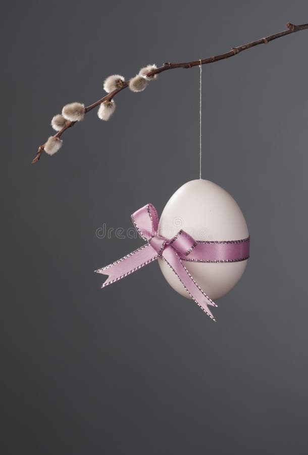 An Easter Egg with a very cute pink bow hanging from a backlit Pussy-Willow Branch on a dark grey background. An Easter Egg with a very cute pink bow hanging from a backlit Pussy-Willow Branch on a dark grey background.