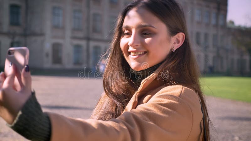 Śliczna uśmiechnięta caucasian dziewczyna stojąca bierze selfies z rozochoconym uśmiechem i trzyma jej telefon z jeden ręką podcz