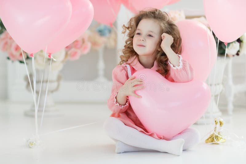 A little girl of 6 years with long curly hair, dressed in a pink dress and white tights, a beautiful smile, sits alone in a large bright room with lots of pink balloons in the shape of a heart. Valentine`s Day and party celebration. A little girl of 6 years with long curly hair, dressed in a pink dress and white tights, a beautiful smile, sits alone in a large bright room with lots of pink balloons in the shape of a heart. Valentine`s Day and party celebration.