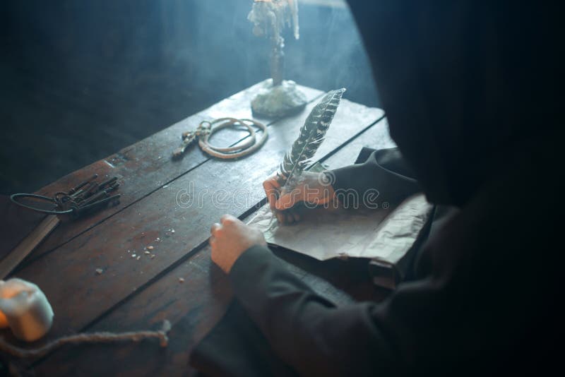 Medieval monk sitting at the table and writes with a goose feather, top view, secret scripture. Mysterious friar in dark cape. Mystery and spirituality. Medieval monk sitting at the table and writes with a goose feather, top view, secret scripture. Mysterious friar in dark cape. Mystery and spirituality