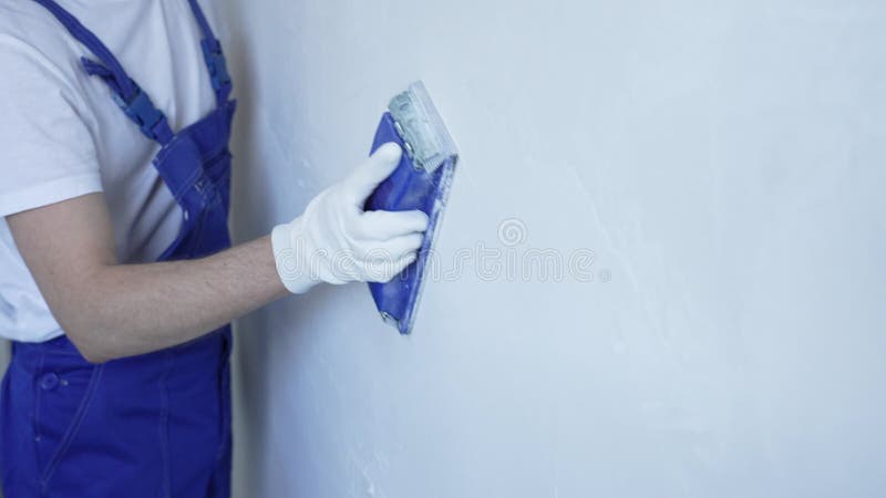 Άγνωστος άνδρας οικοδόμος με προστατευτικά λευκά γάντια και μπλε καλύμματα οικοδομών λειαίνει λευκό τοίχο φιλμ μικρού μήκους