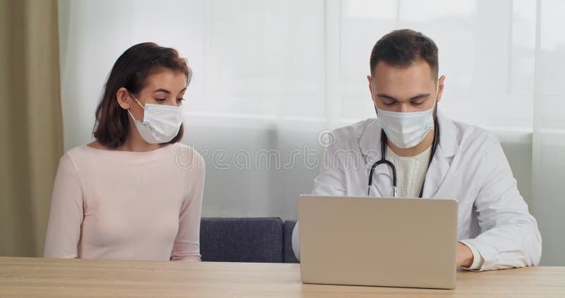 άνδρας γιατρός με ιατρική στολή κάθεται στο τραπέζι σε νοσοκομείο δέχεται συμβουλές γυναίκα ασθενής με ιατρική μάσκα