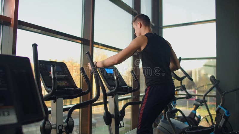 Άνδρας γυμναστικής με πορτραίτο ζεσταίνεται πριν προπονηθεί σε ελλειπτικό σταυρό εκπαιδευτή σε γυμναστήριο Στενοί άνδρες φιλμ μικρού μήκους