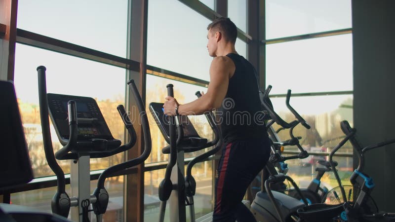 Άνδρας γυμναστικής με πορτραίτο ζεσταίνεται πριν προπονηθεί σε ελλειπτικό σταυρό εκπαιδευτή σε γυμναστήριο Στενοί άνδρες απόθεμα βίντεο