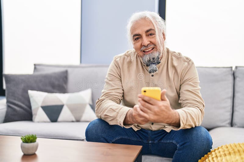 άνδρας με γκριμάτσα μεσαίας ηλικίας με smartphone που κάθεται στον καναπέ στο σπίτι στοκ φωτογραφία με δικαίωμα ελεύθερης χρήσης