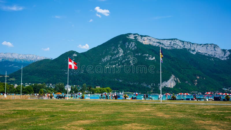Άνθρωποι γύρω από τη λίμνη του Annecy riverbank με Savoie τη σημαία περιοχών και τα βουνά Άλπεων στο υπόβαθρο στοκ φωτογραφία με δικαίωμα ελεύθερης χρήσης Άνθρωποι γύρω από τη λίμνη του Annecy riverbank με Savoie τη σημαία περιοχών και τα βουνά Άλπεων στο υπόβαθρο στοκ φωτογραφία με δικαίωμα ελεύθερης χρήσης