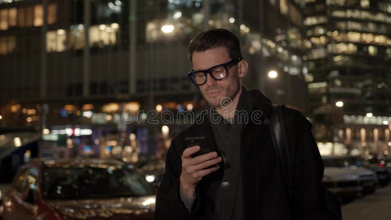 άντρας με γυαλιά με τηλέφωνο στο παρασκήνιο των επαγγελματικών κτιρίων τη νύχτα