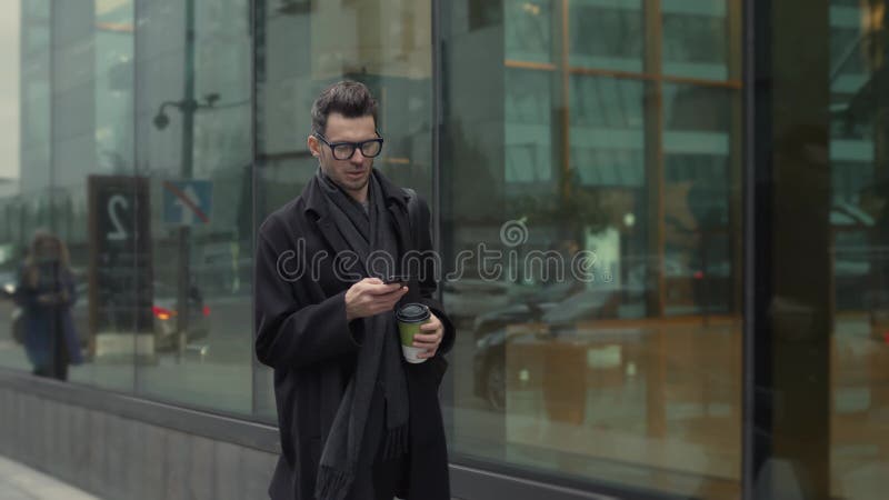 άντρας με μαύρα μάτια και γυαλιά με τηλέφωνο να περπατά στο δρόμο με καφέ