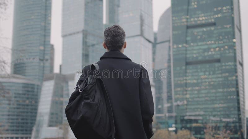 άντρας με παλτό περπατάει προς τα κτίρια επιχειρήσεων αργός ουρανοξύστης