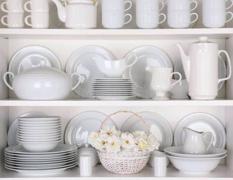 Closeup of white plates and dinnerware in a cupboard. A basket of white roses is centered on the bottom shelf. Items include, plates, coffee cups, saucers, soup tureen, tea pot, and gravy boats. Closeup of white plates and dinnerware in a cupboard. A basket of white roses is centered on the bottom shelf. Items include, plates, coffee cups, saucers, soup tureen, tea pot, and gravy boats.