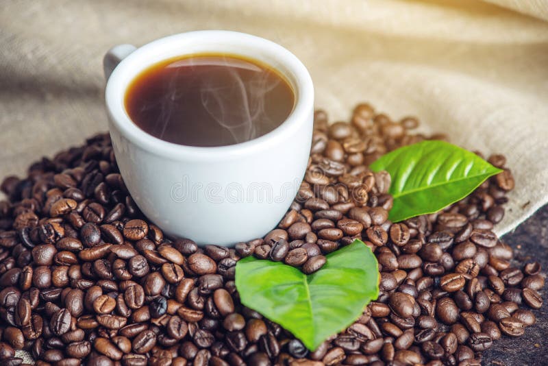 White black espresso Cup with a pile of coffee beans and green leaves in bag on white linen background. The concept of the production of fresh aromatic drink. White black espresso Cup with a pile of coffee beans and green leaves in bag on white linen background. The concept of the production of fresh aromatic drink.