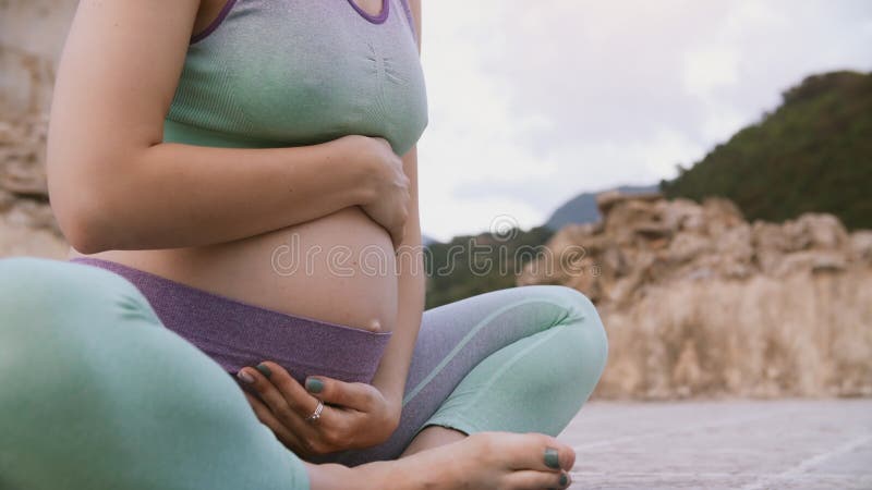Έγκυος κοιλιά με τα χέρια σε το Γυναίκα που κάνει τη συνεδρίαση γιόγκας στο λωτό υπαίθρια