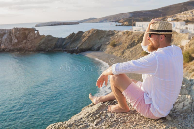 Ένα γενειοφόρο άτομο hipster στις διακοπές κάθεται σε μια απότομη ακτή και εξετάζει την απόσταση στοκ φωτογραφία με δικαίωμα ελεύθερης χρήσης Ένα γενειοφόρο άτομο hipster στις διακοπές κάθεται σε μια απότομη ακτή και εξετάζει την απόσταση στοκ φωτογραφία με δικαίωμα ελεύθερης χρήσης