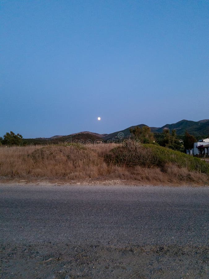 Ένα διπλό τοπίο του λυκόφωτος παρουσιάζει ένα μακρινό φεγγάρι πάνω από τους κυλιόμενους λόφους και μια ήσυχη όψη στην άκρη του δρό στοκ εικόνες