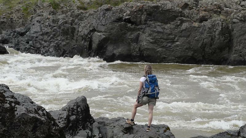 Ένα κορίτσι με ένα σακίδιο πλάτης στέκεται στις όχθεις ενός ποταμού βουνών