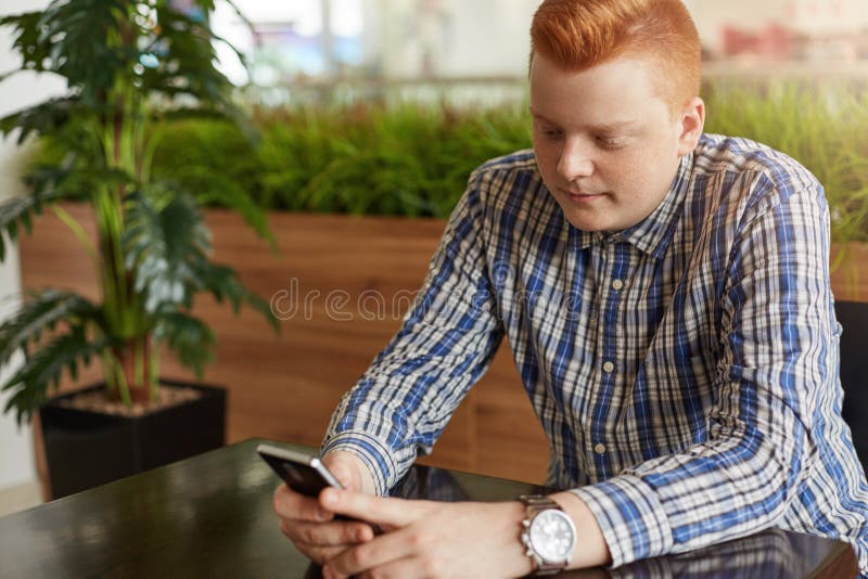 Ένα μοντέρνο redhead άτομο έντυσε στην ελεγχμένη συνεδρίαση πουκάμισων στο άνετο εστιατόριο κοντά στον πράσινο φοίνικα χρησιμοποι στοκ εικόνες Ένα μοντέρνο redhead άτομο έντυσε στην ελεγχμένη συνεδρίαση πουκάμισων στο άνετο εστιατόριο κοντά στον πράσινο φοίνικα χρησιμοποι στοκ εικόνες