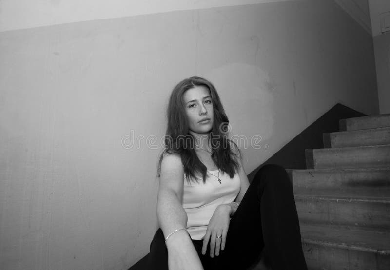 Ένα νεαρό όμορφο κορίτσι με χαλαρά μαλλιά σε ένα μπλουζάκι και παντελόνια κάθεται στην είσοδο του σπιτιού στην προσγείωση με μια στοκ φωτογραφία με δικαίωμα ελεύθερης χρήσης Ένα νεαρό όμορφο κορίτσι με χαλαρά μαλλιά σε ένα μπλουζάκι και παντελόνια κάθεται στην είσοδο του σπιτιού στην προσγείωση με μια στοκ φωτογραφία με δικαίωμα ελεύθερης χρήσης