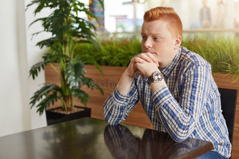 Ένα λοξά πορτρέτο του redhead τύπου που φορά την ελεγχμένη συνεδρίαση πουκάμισων και ρολογιών στον καφέ στον ξύλινο πίνακα κοντά στοκ φωτογραφίες Ένα λοξά πορτρέτο του redhead τύπου που φορά την ελεγχμένη συνεδρίαση πουκάμισων και ρολογιών στον καφέ στον ξύλινο πίνακα κοντά στοκ φωτογραφίες