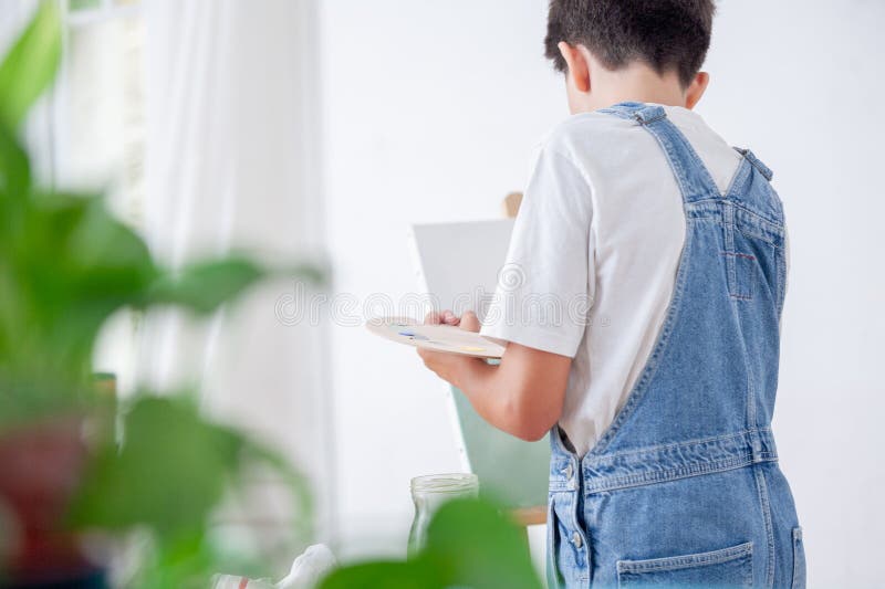 ένα παιδί με μπλε φόρμες ζωγραφίζει μια εικόνα. κρατά πινέλο και παλέτα. στοκ φωτογραφία με δικαίωμα ελεύθερης χρήσης