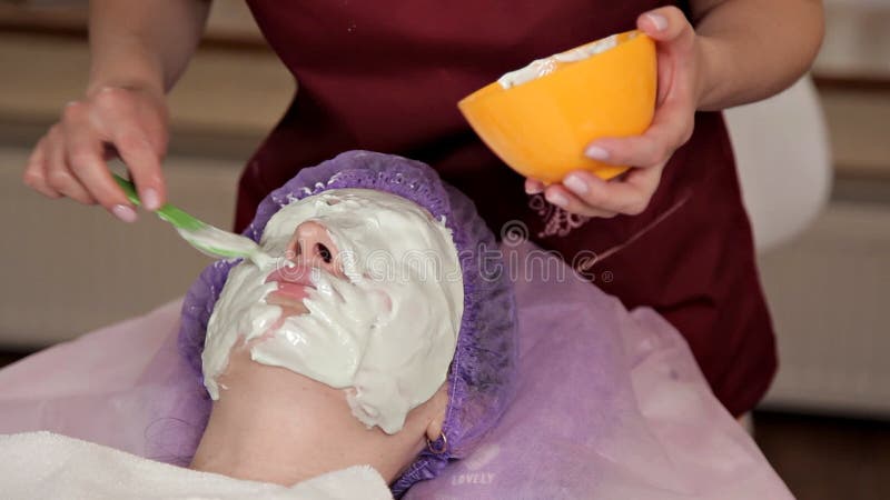 Ένα πεπειραμένο cosmetologist προετοιμάζει μια υγιή μάσκα για τον πελάτη Παίρνει επάνω και αγγίζει το πρόσωπο με μια βούρτσα ?