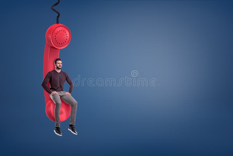 A casual looking bearded man sits on a giant red retro phone receiver hanging from a cord. Call center worker. Waiting for important call. Priority message. A casual looking bearded man sits on a giant red retro phone receiver hanging from a cord. Call center worker. Waiting for important call. Priority message.