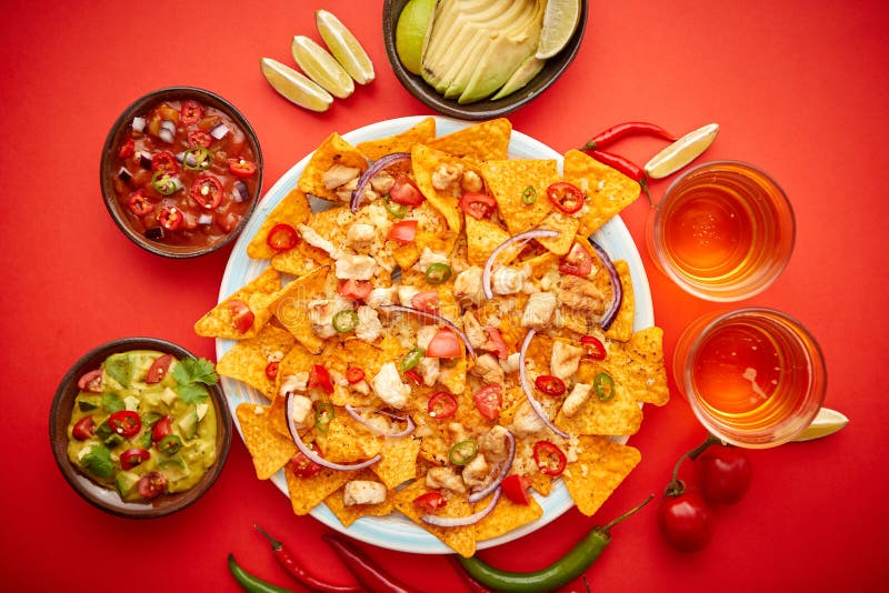 A plate of delicious tortilla nachos with melted cheese sauce, grilled chicken, jalapeno peppers, red onion, tomato, guacamole dip and spicy salsa. With cold sparkling beer. Placed on red background. A plate of delicious tortilla nachos with melted cheese sauce, grilled chicken, jalapeno peppers, red onion, tomato, guacamole dip and spicy salsa. With cold sparkling beer. Placed on red background