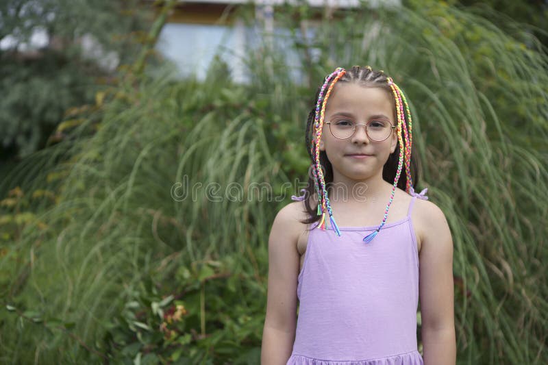 Ένα χαριτωμένο χαμογελαστό και θετικό κορίτσι με γυαλιά και ένα φόρεμα με πολύχρωμα μικρά πλεξούδα σε μια αυλή του καλοκαιριού στοκ φωτογραφία με δικαίωμα ελεύθερης χρήσης